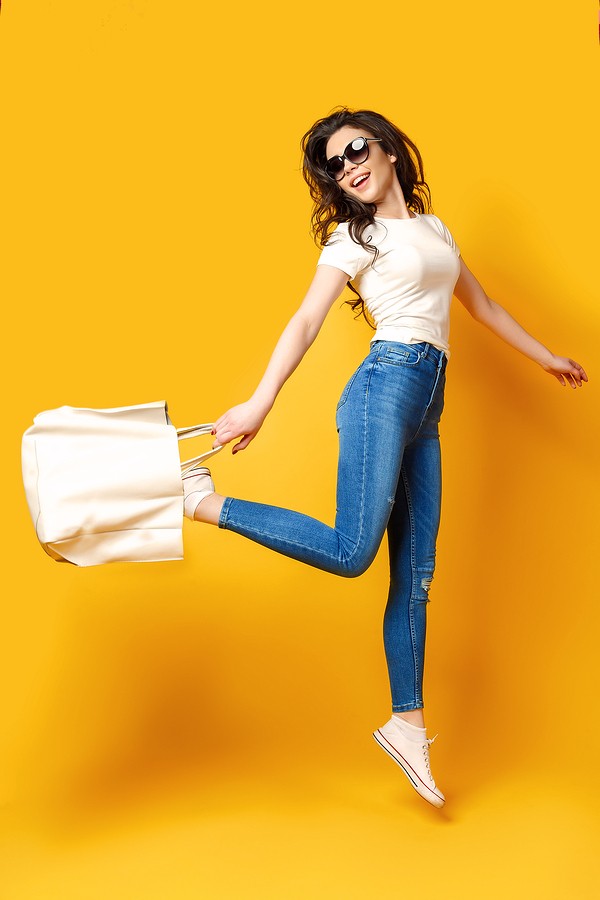 Beautiful Young Woman In Sunglasses, White Shirt, Blue Jeans Posing, Jumping With Bag On The Yellow 